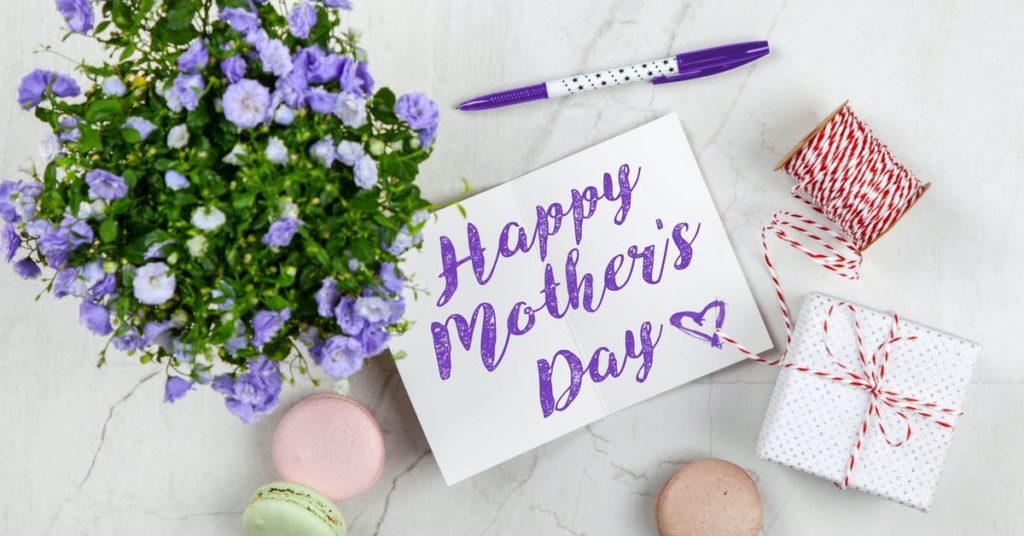 Bouquet of purple flowers, Mother's Day card with purple greeting, small box wrapped in white gift wrapping and red twine, and macarons on a marble surface