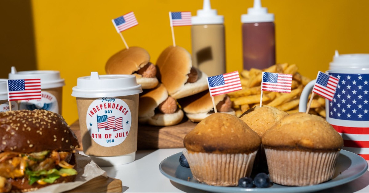 Close-up of a Fourth of July cookout with muffins, hot dogs, hamburger, & fries; all foods have American flag toothpick decoration; yellow background